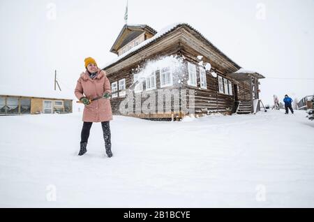 Febbraio 2020 - Factory Village. Bella ragazza pulisce la neve vicino a una grande casa di villaggio di legno. Inverno nel villaggio russo Foto Stock