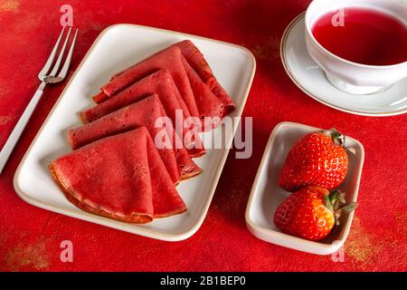 Avvicinati a deliziosi pancake sottili di fragole rosse fatti in casa con fragole fresche e tè alla frutta rossa. Vista dall'alto, primo piano su sfondo rosso Foto Stock