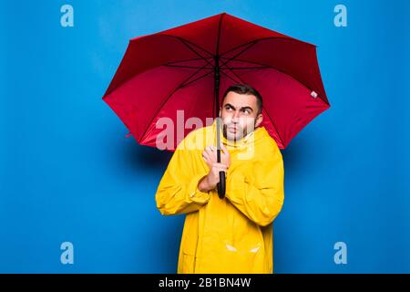 Giovane bell'uomo portacolori in impermeabile giallo con copertura ombrello rosso dalla pioggia freddo su sfondo blu Foto Stock