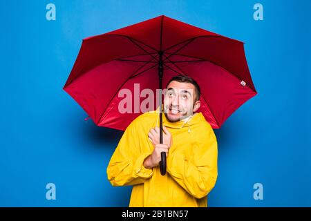 Giovane bell'uomo portacolori in impermeabile giallo con copertura ombrello rosso dalla pioggia freddo su sfondo blu Foto Stock