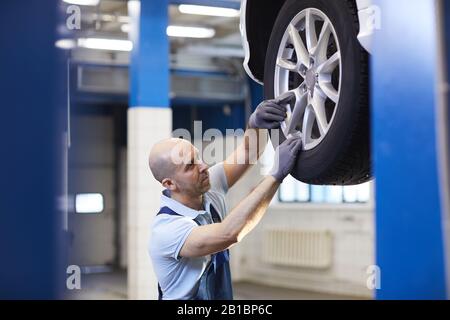 Ritratto in alta sezione del meccanico dell'automobile del bald che controlla le ruote del veicolo sull'elevatore dell'automobile durante l'ispezione nell'officina del garage, spazio della copia Foto Stock