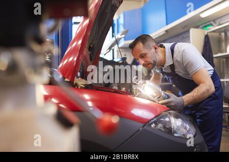 Vista laterale ritratto di meccanico di automobile con bearded che guarda sotto il cofano durante l'ispezione del veicolo in officina, copia spazio Foto Stock