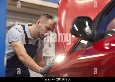 Vista laterale ritratto di meccanico di automobile con bearded che guarda sotto il cofano durante l'ispezione del veicolo in officina, copia spazio Foto Stock