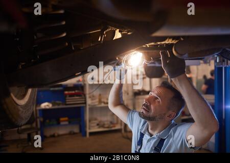 Vista in sezione alta del meccanico maturo che guarda sotto la vettura sul sollevatore e la luce della lampada durante l'ispezione in officina per la riparazione automatica, copiare spazio Foto Stock