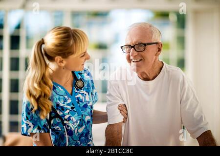 Un'infermiera amichevole giovane di riposo a casa parla con il suo paziente anziano mentre lo aiuta a salire dal letto. Foto Stock