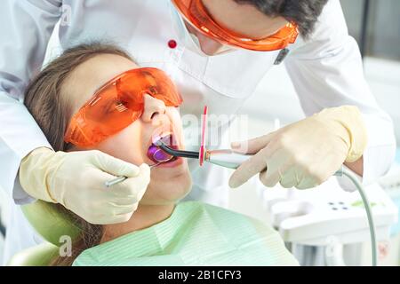 Ragazza bambino al medico. Il dentista posiziona un riempimento su un dente con la lampada di polimerizzazione dentale in cavità orale. Su sfondo clinico Foto Stock