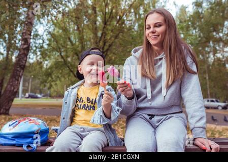 Donna mamma con suo figlio, ragazzino mangia gelato rosa sul bastone. In estate città, abbigliamento casual, felice gioisce, ha divertimento deliziato con delizioso ghiaccio Foto Stock