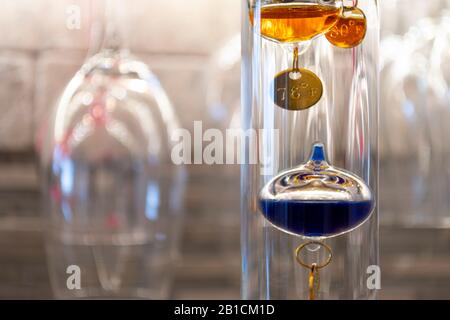 Un termometro Galileo colorato, in vetro e riempito di liquido misura la temperatura ambiente interna in una cantina con bicchieri da vino appesi dietro. Foto Stock
