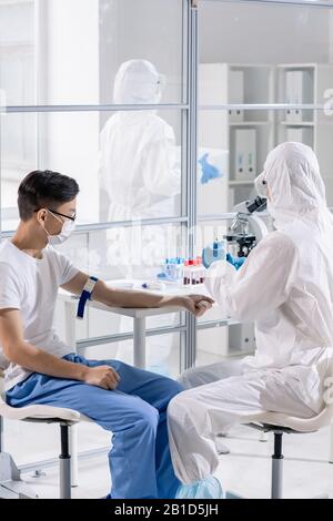 Medico in tuta protettiva che prende sangue di pazienti asiatici di coronavirus durante la ricerca medica in laboratorio Foto Stock