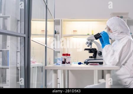 Vista laterale dello scienziato medico in maschera protettiva al microscopio mentre studia il coronavirus pericoloso in laboratorio Foto Stock