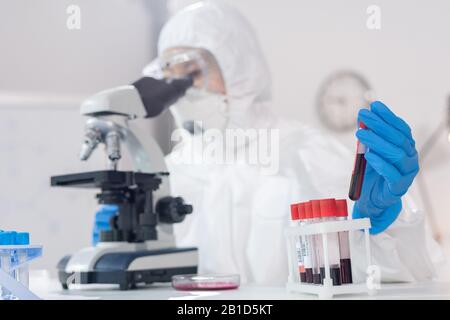 Primo piano del tecnico di laboratorio nella tuta protettiva che esamina i campioni di sangue al microscopio in laboratorio Foto Stock