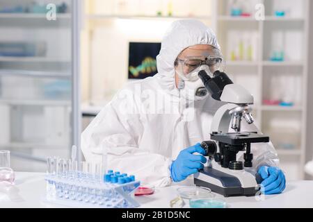 Scienziato medico asiatico in respiratore e occhiali di sicurezza utilizzando il microscopio per la ricerca di campioni di sangue infetti in laboratorio Foto Stock