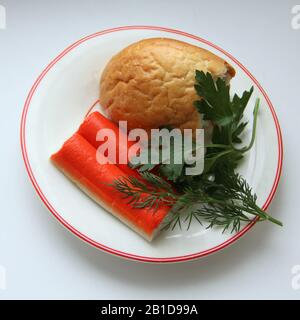 Ancora vita di un pezzo di pane bianco, granchio bastoni e prezzemolo su un piatto bianco con un bordo rosso. Foto di scorta per il Web e la stampa, il walpaper e lo sfondo. Foto Stock