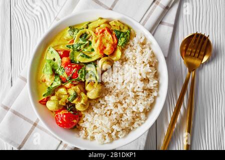 Pasto vegano al cocco e al curry di funghi con spinaci freschi, pomodori ciliegini serviti con riso su un piatto bianco con posate dorate su un tabl di legno bianco Foto Stock