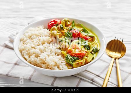 Riso appiccicoso con curry di cocco giallo con spinaci freschi, pomodori ciliegini serviti su un piatto bianco con posate dorate su uno sfondo di legno bianco, ho Foto Stock