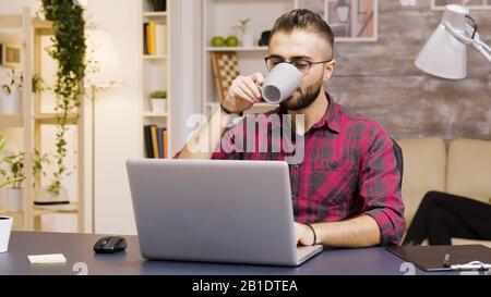 Bel freelancer aprire il suo portatile per iniziare a lavorare in salotto. Prendere un sorso di caffè. Foto Stock