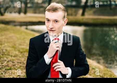 giovane ragazzo in abito nero raddrizza la sua cravatta rossa. uomo biondo con una penna nelle sue mani Foto Stock