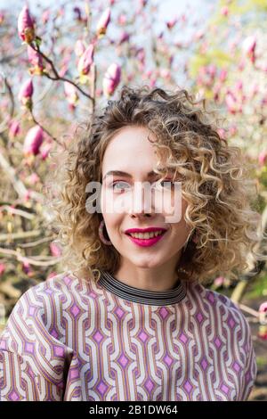 Ritratto di ragazza bella bionda con capelli ricci lunghi. Donna passeggiate nel giardino di fioritura magnolia rosa Foto Stock