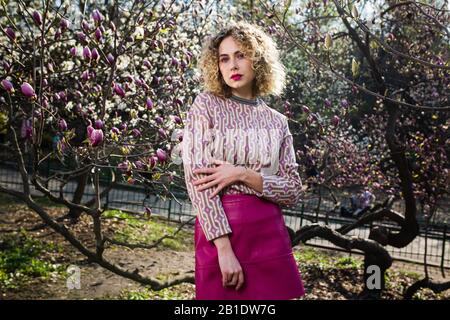 Ritratto di ragazza bella bionda con capelli ricci lunghi. Donna passeggiate nel giardino di fioritura magnolia rosa Foto Stock