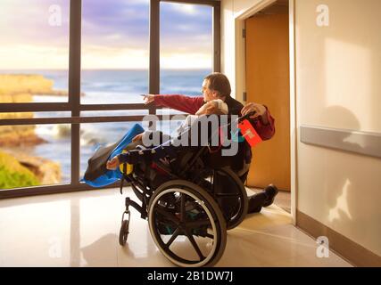 Padre inginocchiato accanto a figlio disabile in sedia a rotelle che guarda fuori finestra ospedale in oceano e scogliere all'aperto. Recentemente il bambino ha avuto un intervento chirurgico alla testa. Foto Stock
