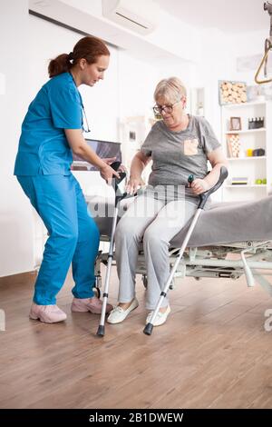 Infermiere che aiuta la vecchia donna a sedersi a letto in casa di cura dopo aver camminato con stampelle. Foto Stock