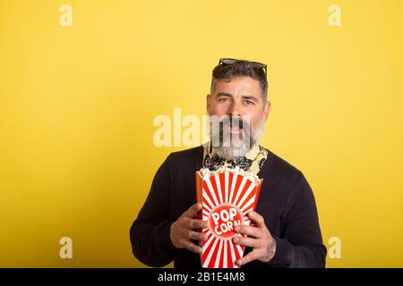 Ritratto di uomo attraente con barba bianca che guarda sorridendo verso la fotocamera con una scatola di popcorn su sfondo giallo. Foto Stock