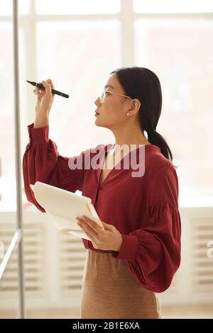Ritratto di vista laterale di moderna donna d'affari asiatica che scrive su parete di vetro mentre progettano il progetto in ufficio, spazio della copia Foto Stock