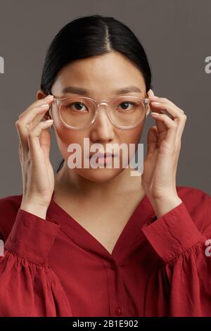 Testa e spalle ritratto di elegante donna asiatica mettere su occhiali mentre in piedi su sfondo grigio in studio Foto Stock