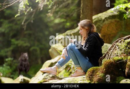 Giovane ragazza seduta nei boschi guardando il telefono e l'orso cammina sul lato sullo sfondo Foto Stock