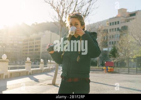 Bella ragazza con trecce tiene tazza di caffè e utilizza macchina fotografica nel centro della città con cappotto soffusa Foto Stock