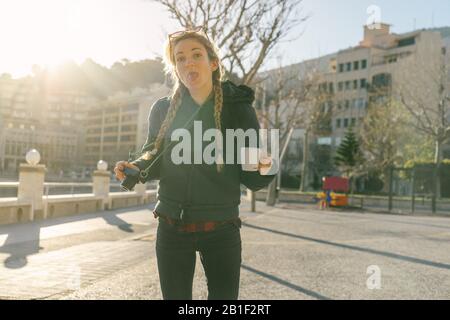 Bella ragazza con trecce tiene tazza di caffè e utilizza macchina fotografica nel centro della città con cappotto soffusa Foto Stock