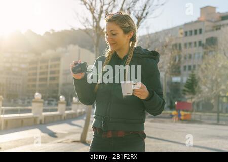 Bella ragazza con trecce tiene tazza di caffè e utilizza macchina fotografica nel centro della città con cappotto soffusa Foto Stock