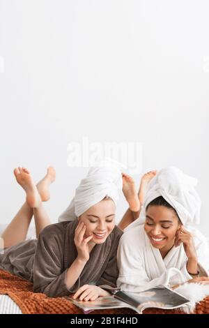 Due graziose Signore in accappatoi che posano sul letto, riviste di lettura Foto Stock