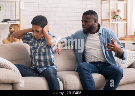 Piccolo afro ragazzo che copre le orecchie per non ascoltare arrabbiato padre scolding Foto Stock