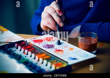 La mano sporca dell'artista in abiti blu contiene una spazzola che mescola vernici acquerello su una tavolozza che giace su un tavolo di legno, e c'è un vetro Foto Stock