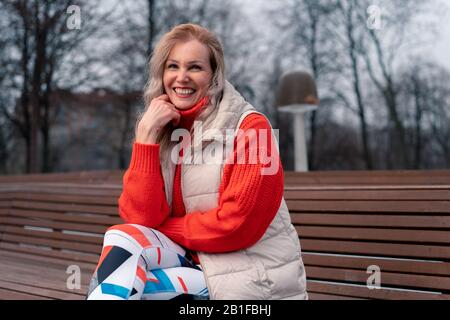 Donna Bionda Matura Sorridente E Seduto Su Bench Nel Parco Moderno Dopo L'Esercizio Attivo All'Aperto. Stile Di Vita Delle Persone Anziane. Sport E Relaxatio Foto Stock