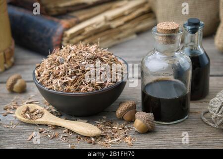 Corteccia sana di quercia in recipiente di ceramica nera, bottiglia per infusione e libri antichi su tavolo di legno. Medicina di erbe. Foto Stock