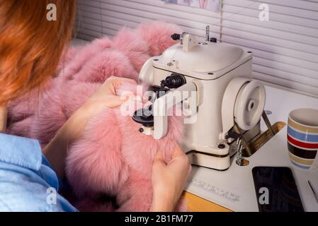 Master professionale per la produzione di capi di abbigliamento da pelliccia. Donna degno alla macchina da cucire cuce i vestiti da pelliccia rosa. Il processo di cucitura Foto Stock