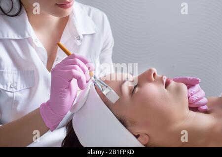 primo piano, le mani di un medico cosmetologo professionista, utilizzando uno strumento, esegue l'applicazione di crema e olio sulla pelle del viso di una giovane pat Foto Stock