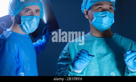 Ritratto di due chirurghi in abiti sterili, cappelli e maschere. Un chirurgo, che si prepara ad operare, tiene un bisturi con un supporto per l'ago nelle sue mani, anoth Foto Stock