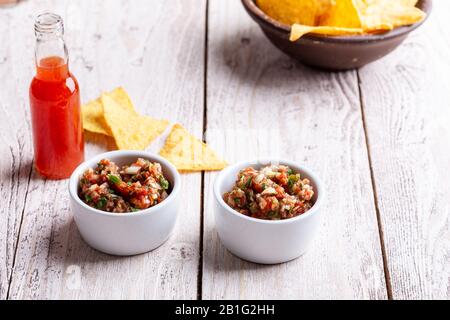 Due ciotole piene di salsa, salsa calda al peperoncino rosso bottiglia e tortilla chip su rustico tavolo di legno Foto Stock