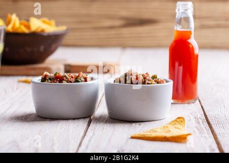 Due ciotole piene di salsa, salsa calda al peperoncino rosso bottiglia e tortilla chip su rustico tavolo di legno Foto Stock