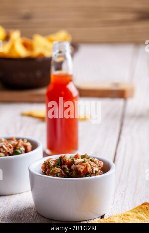 Due ciotole piene di salsa, salsa calda al peperoncino rosso bottiglia e tortilla chip su rustico tavolo di legno Foto Stock