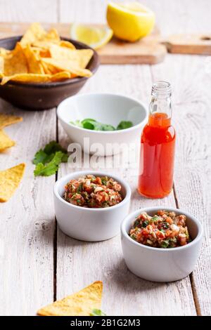 Due ciotole piene di salsa, salsa calda al peperoncino rosso bottiglia e tortilla chip su rustico tavolo di legno Foto Stock