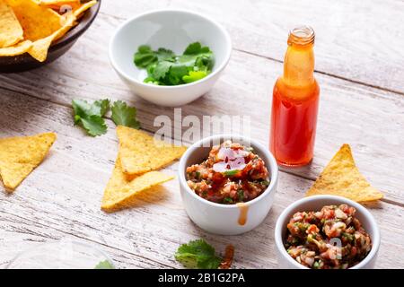 Due ciotole piene di salsa, salsa calda al peperoncino rosso bottiglia e tortilla patatine su tavola rustica in legno, sause che è aggiunto ad una delle ciotole Foto Stock