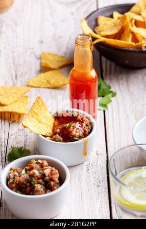 Due ciotole piene di salsa, salsa calda al peperoncino rosso bottiglia e tortilla patatine su tavola rustica in legno, sause che è aggiunto ad una delle ciotole Foto Stock