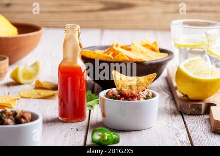 Due ciotole piene di salsa, salsa calda al peperoncino rosso bottiglia e tortilla patatine su tavola rustica in legno, sause che è aggiunto ad una delle ciotole Foto Stock