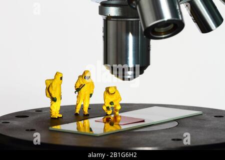 Immagine concettuale della figura in miniatura persone che indossano abiti Hazmat guardando un campione di sangue falso sotto un microscopio Foto Stock