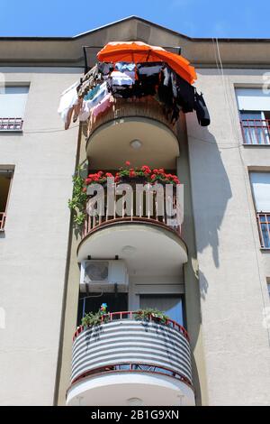 Tre livelli di balconi arrotondati fissati con recinto di metallo rosso riempito di fiori e vestiti che si asciugano sul lato del vecchio edificio appartamento con chiaro Foto Stock