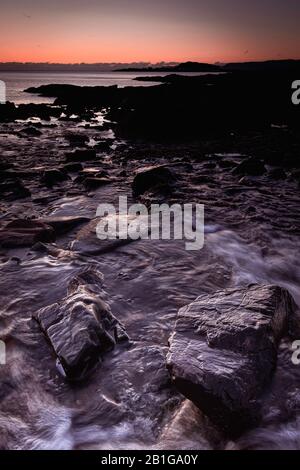 Tramonto sulla spiaggia rocciosa panoramica con bassa marea a Larach na Gaihbre su Argyll e Bute costa della Scozia, UK.Water che scorre tra rocce bagnate cattura sera Foto Stock